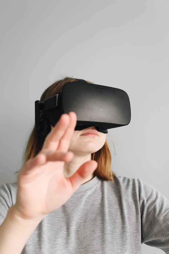 A woman in a gray shirt explores virtual reality with a VR headset indoors against a white wall.
